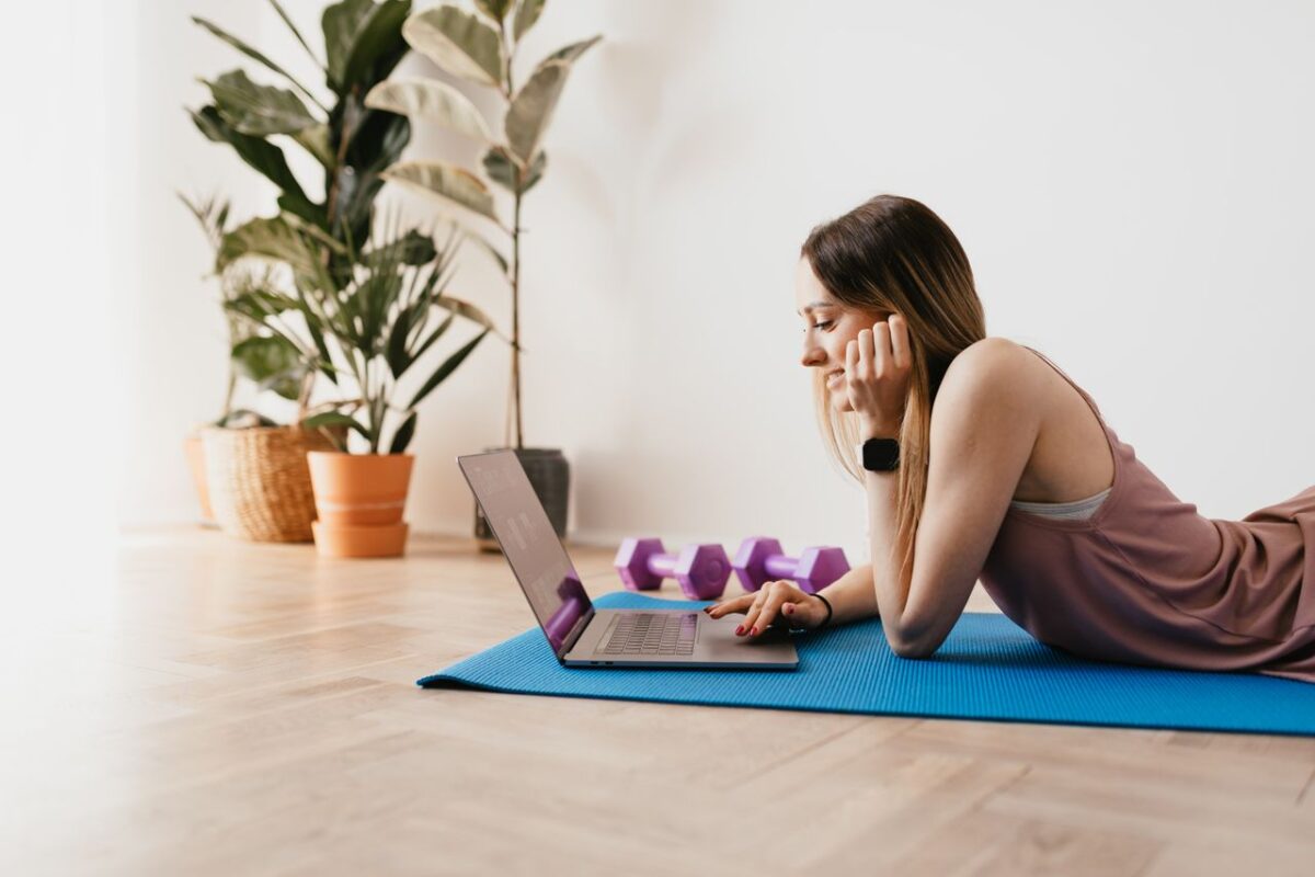 Side view of young cheerful slim female with long hair surfing laptop while lying on blue yoga mat during workout break