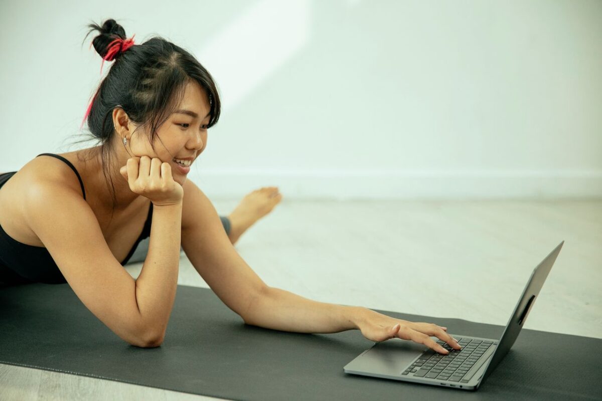 Smiling woman using laptop while stretching leg on mat with hand at chin in light apartment